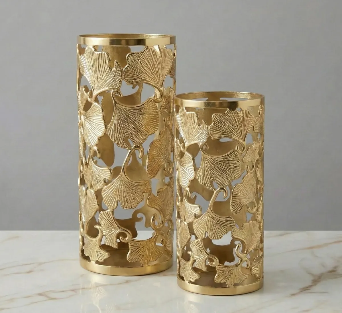 Set of two gold cylindrical metal vases with cutout leaf design and solid rims.